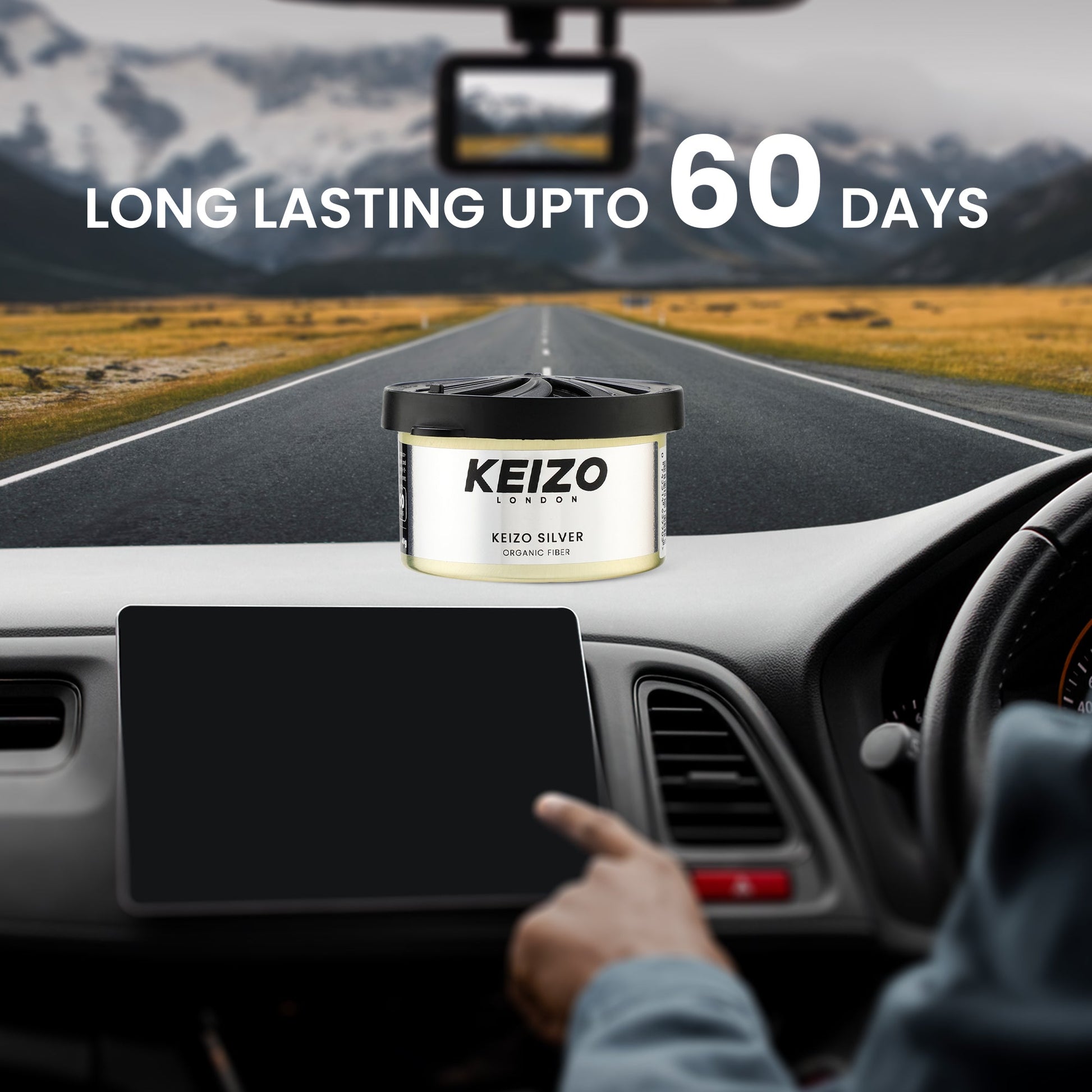 A hand points to a car dashboard with a Keizo London Silver 40g Car Perfume by Keizo, promising up to 60 days of freshness. Through the windshield, a scenic road and mountains complete the view.